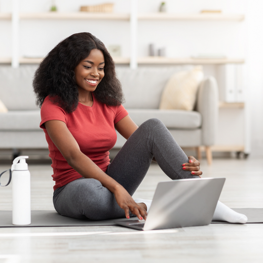 An African-American woman sitting comfortably on the floor in front of a laptop, engaged in a virtual coaching session with a focused expression.